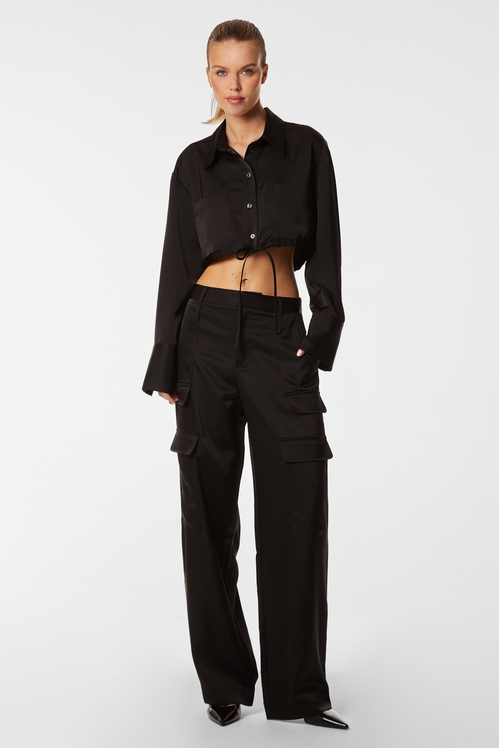 A woman stands against a plain white background, wearing a black, long-sleeve crop top shirt with large cuffs and the Milan Satin Cargo Pant - Black, featuring mid-rise and multiple pockets. She has her hair pulled back and poses with one hand in a pocket, exuding a casual yet stylish look.