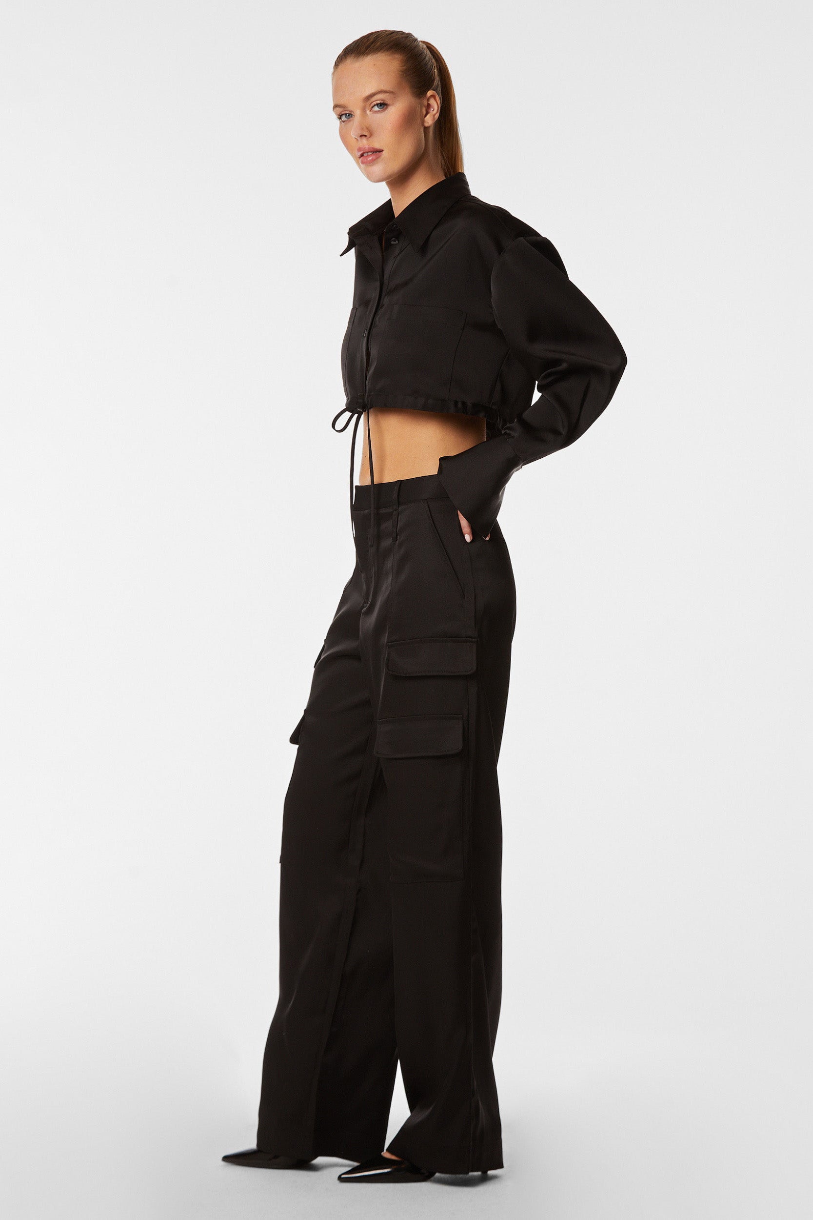 A woman with blonde hair in a ponytail is wearing a black cropped jacket and the Milan Satin Cargo Pant - Black, featuring mid-rise high-waist and side pockets, made from luxurious fabric. She stands against a white background, looking slightly to her left with her hands in her pockets.