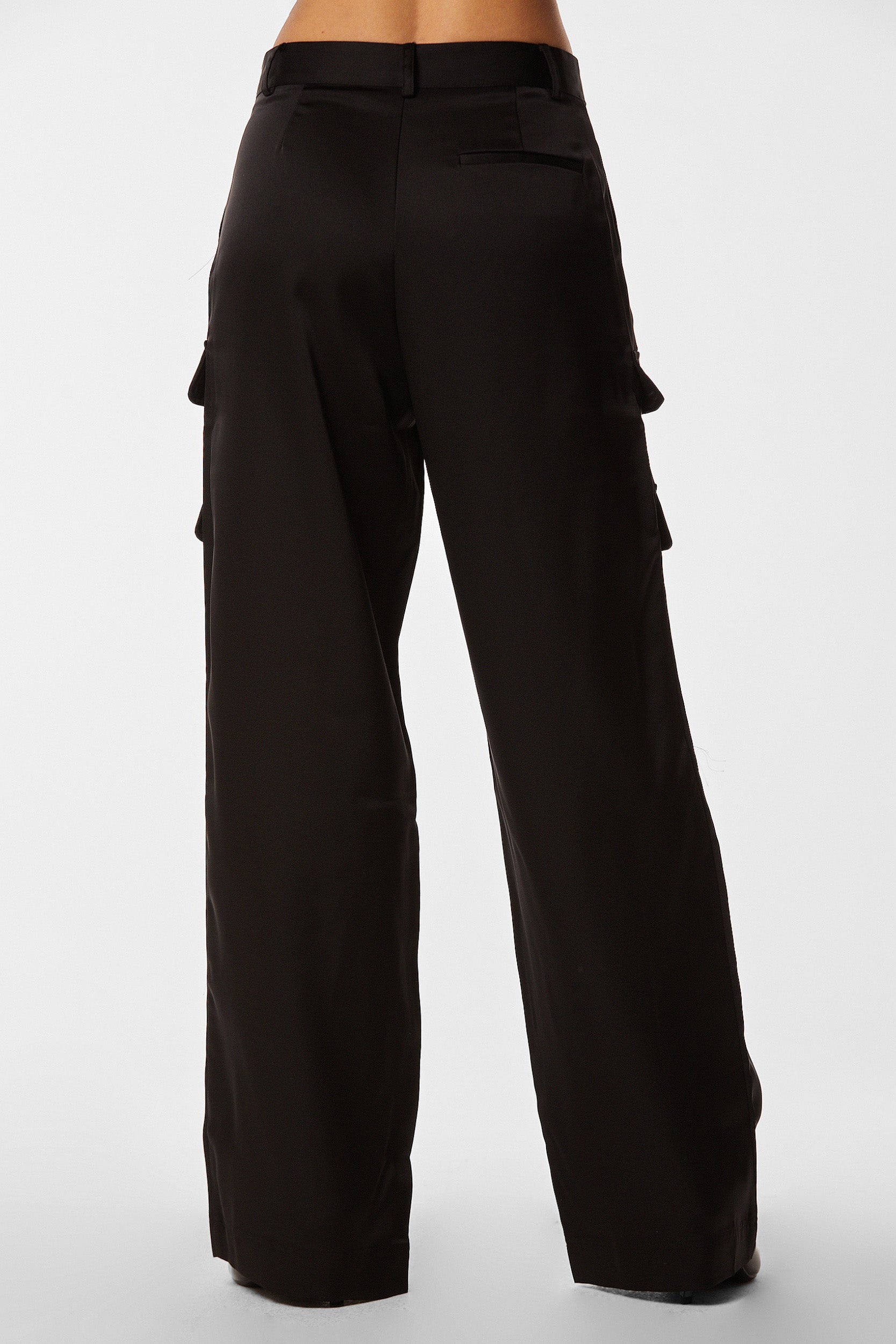 A person is wearing the Milan Satin Cargo Pant in black, high-waisted with a wide-leg design, shown from the back. These versatile trousers feature side pockets with flaps and have a relaxed fit. Crafted from luxurious satin fabric, the plain white background highlights the garment beautifully.