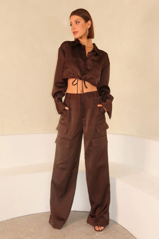 A person stands against a neutral background, wearing an espresso satin-like outfit. The attire includes a cropped shirt with long sleeves and drawstring detail, and mid-rise, wide-legged Milan Satin Cargo Pants with multiple pockets, paired with open-toe black heels.