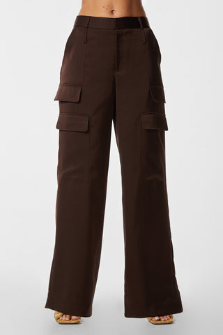 A person is wearing the Milan Satin Cargo Pant in Espresso from Noli Yoga, featuring multiple pocket flaps on the front. The versatile, high-waisted trousers are wide-legged and paired with tan high-heeled sandals. The background is plain white.