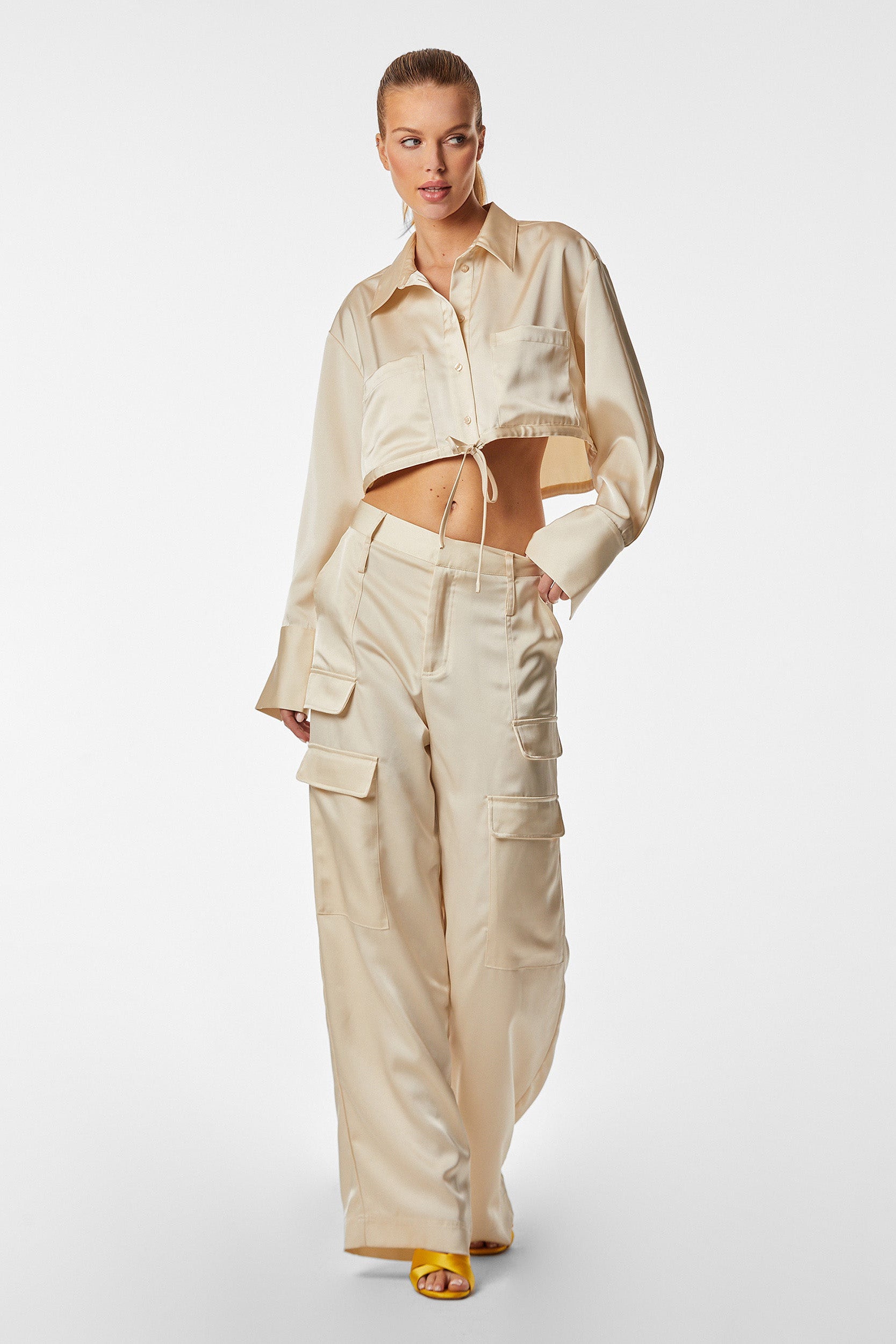 A person wearing the Milan Satin Cargo Pant in Pearl paired with an off-white, long-sleeve, cropped button-up shirt made from satin-like fabric. They have one hand in their pocket and are walking towards the camera, also sporting yellow open-toe heels.