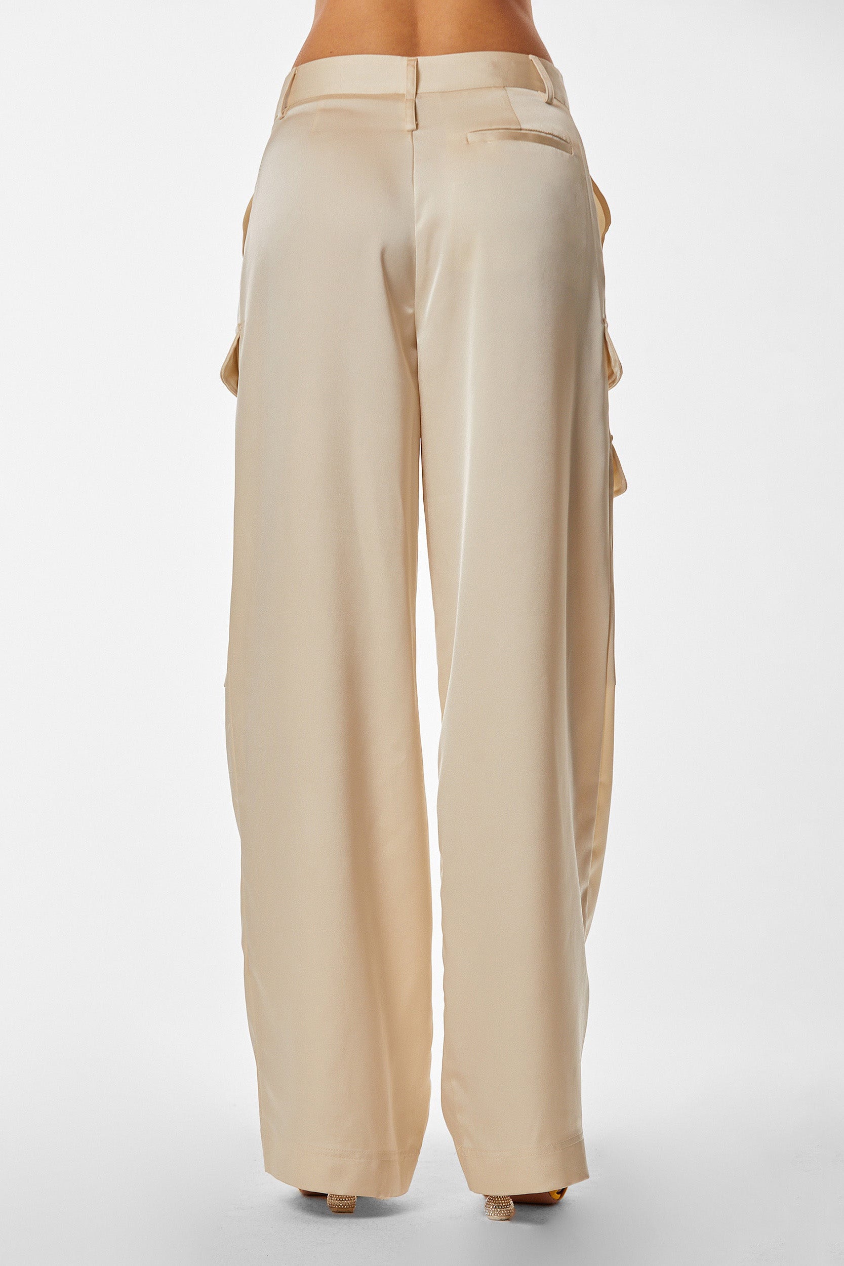 A person wearing high-waisted, wide-leg Milan Satin Cargo Pant in Pearl is shown from the back. The pants are made of a smooth satin-like fabric and have a relaxed fit with subtle pleats and pockets on the back. The background is a plain white studio setting.