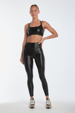 A person models the Liquid Jolie Bra - Black Gloss, crafted from moisture-wicking fabric, paired with matching leggings against a plain white background. They wear black and white sneakers, with one hand on their hip and the other relaxed at their side.