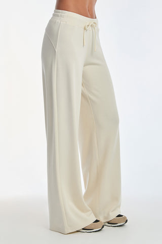 A person models the Daphne Pant - Pearl, featuring a luxurious cream-colored modal blend with wide legs and a drawstring waist. They pair it with black and white sneakers against a plain white background, exuding effortless comfort.