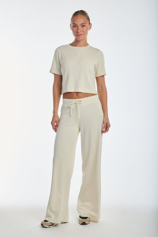 Dressed in a luxurious light beige ensemble of a short-sleeve, cropped top and wide-leg modal blend drawstring pants, the person wears black and beige sneakers against a plain white background, highlighting the comfort of the Daphne Pant - Pearl.