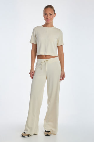 A person wears the Daphne Soft T-Shirt in Pearl, a light beige short-sleeve cropped top, with matching wide-leg pants. They complete their outfit with black and white sneakers against a white background.