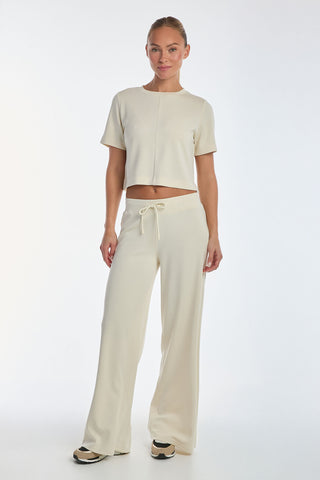 A person embodies the ultimate on-the-go style in a cream short-sleeve crop top, matching Daphne Pant - Pearl in a soft modal blend, and black and white sneakers, standing against a plain white background.