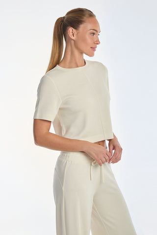 A woman with a ponytail wears the Daphne Soft T-Shirt - Pearl and matching pants in heathered modal. Standing sideways, hands gently by her side, she exudes an effortless silhouette against a soft white background.