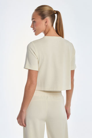 A person with a ponytail wears the Daphne Soft T-Shirt in Pearl, paired with matching pants. The short-sleeve top shows a t-shirt silhouette as they turn to the side, facing left against a plain white background.