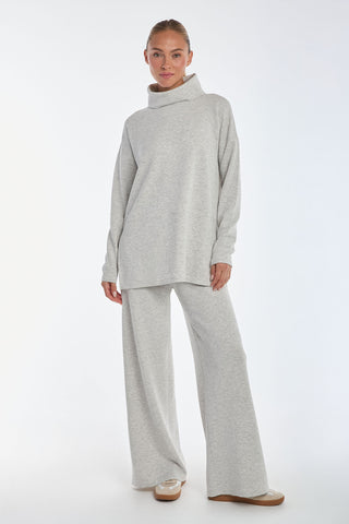 Clad in a light gray oversized turtleneck and Heather Grey Daphne Pant, a person stands against a white backdrop with their hair tied back, brown shoes on, radiating luxurious comfort.