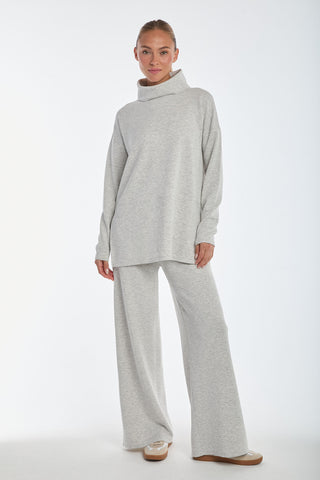 Wearing a loose-fitting, high-collar light gray sweater in a soft modal blend paired with Daphne Pant - Heather Grey, this person strikes a comfortable and casual pose against a white background with their hair pulled back neatly.