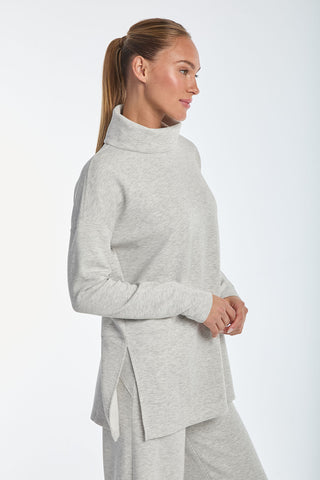 A person with long hair tied back wears the Daphne Top in Heather Grey and matching pants, made from a soft modal blend. They stand sideways with hands clasped, looking forward against a plain background, exuding luxurious comfort.