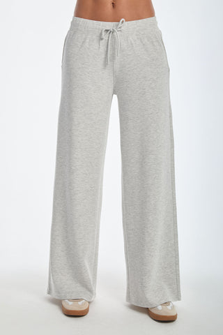 Wearing the Daphne Pant in Heather Grey, featuring wide legs and a soft modal blend with an elastic waistband and drawstring, paired with beige sneakers. This silky, breathable look offers ultimate on-the-go style against a plain white background.