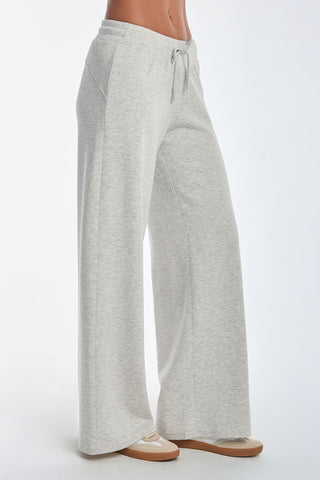 A person wears light gray, wide-leg Daphne Pant in Heather Grey, made from a soft modal blend with a drawstring waistband and pockets, exuding luxurious comfort. They pair it with beige sneakers against a plain white background.