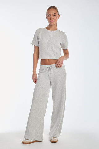 Wearing a Daphne Soft T-Shirt in Heather Grey and matching wide-leg pants, a person stands with relaxed posture against a plain white background, looking towards the camera. The ultra-soft crop top enhances the outfit's comfortable and casual feel.