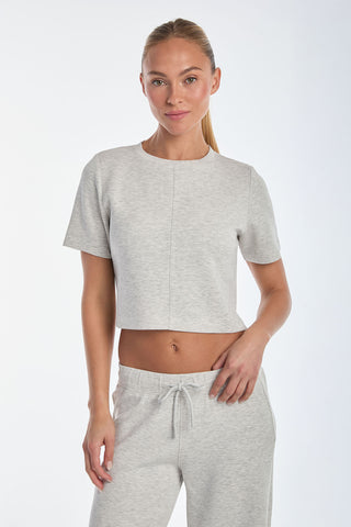 A person in a Daphne Soft T-Shirt - Heather Grey and matching joggers, made from ultra-soft Daphne fabric, poses against a plain white background. With long hair tied back and one hand on their hip, they showcase this must-have staple for effortless style.