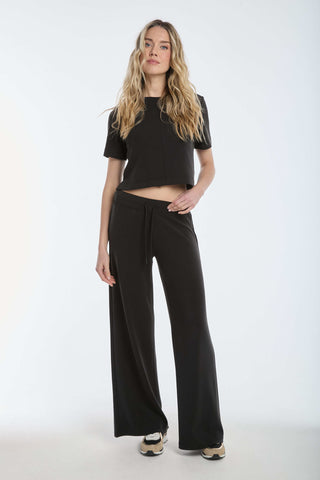 A person with long, wavy hair is wearing a Daphne Soft T-Shirt in black paired with wide-leg pants and sneakers. The scene is set in a plain white studio.