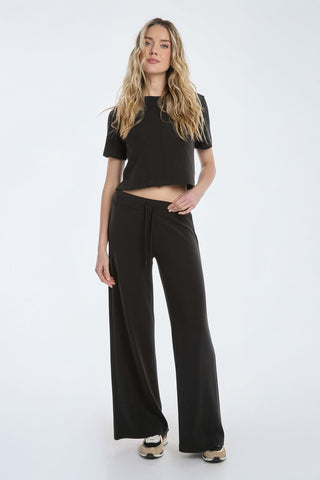 A woman with long, wavy blonde hair dons the Daphne Soft T-Shirt in black and wide-leg pants, standing against a white background. She pairs the look with beige and black sneakers, maintaining a relaxed posture with one hand on her hip.