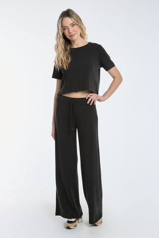 A person with long wavy hair stands against a white background, smiling slightly. They wear the Daphne Soft T-Shirt - Black, a must-have staple crafted from lightweight modal, paired with wide-leg pants. One hand is on their hip while the other rests relaxed by their side.