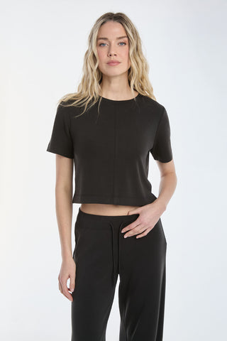 Someone with long, wavy hair is wearing a Daphne Soft T-Shirt in black and matching pants, standing against a plain white background, looking forward with one hand by their side.