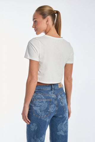 A woman in a white Diana Bow T-Shirt and high-waisted Rosalie Jeans with floral prints on the back pockets stands, looking left. Her ponytail features a satin bow, set against a plain white background.