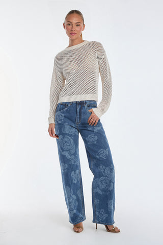 Against a plain backdrop, a person models the Rose High Rise Jean featuring floral patterns. They're paired with a mesh knit sweater, one hand casually in their pocket, and completed the look with open-toe heels.