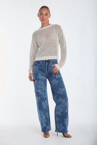 A person stands against a plain background, wearing a textured white sweater, Rose Print High Rise Relaxed Jeans in medium blue, and beige heels. They have one hand in their pocket and gaze forward.