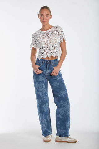 A person stands confidently in a Sadie Lace T-Shirt - White and high-waisted Dahlia Jeans with floral patterns. They wear white sneakers with tan accents, and their hair is pulled back against a plain white background.