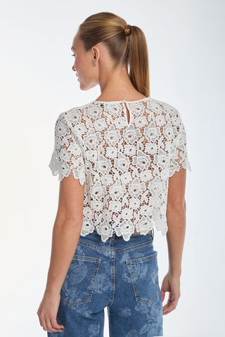 A woman sporting a ponytail wears the Sadie Lace T-Shirt in white, paired with high-waisted jeans. She faces away, showcasing the intricate floral lace design of her top against a plain, light background.