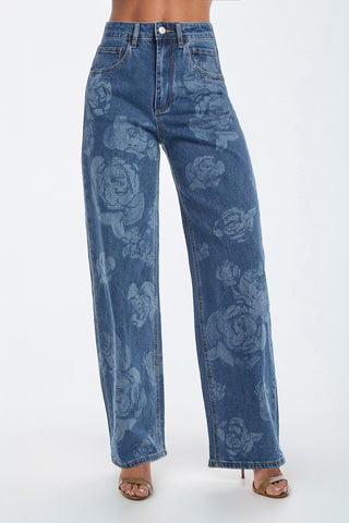 A person in Rose Print High Rise Relaxed Jeans - Medium Blue with a large floral rose pattern, front pockets, and visible seams stands against a plain background, wearing open-toed sandals.