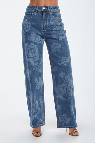 A woman wears the Rose Print High Rise Relaxed Jean in medium blue, showcasing a floral rose design. The jeans feature a wide-leg cut with front and back pockets, paired with light brown heels against a plain white backdrop.