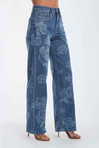 A person wears Rose High Rise Jeans, featuring a floral-patterned, wide-leg design and classic five-pocket style. The look is finished with open-toe heels on a plain white background.