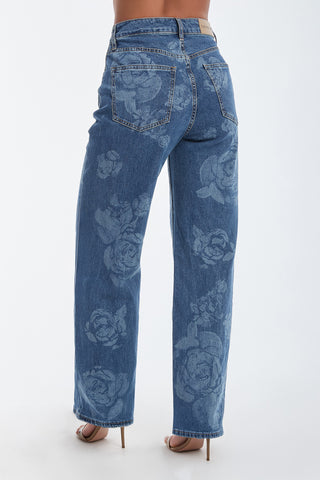 A woman elegantly wears the Rose Print High Rise Relaxed Jean in medium blue, featuring rose blooms on the back pockets and down the legs, paired with beige high-heeled shoes.