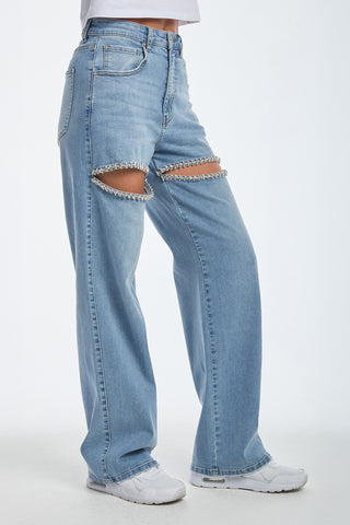 The person is wearing medium blue high-waisted jeans, known as the Ivanka Crystal Jean, with rhinestone slits around the ripped knees, paired with a white top and sneakers. These jeans feature a relaxed fit and straight-leg design, adding sparkle to the casual look.