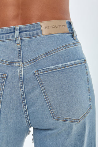 Close-up of someone in Medium Blue Ivanka Crystal Jeans featuring visible back pockets and a brown leather patch labeled "THE NOLI SHOP" on the waistband. Rhinestone slits add sparkle, with the white background highlighting the jeans' exquisite details.