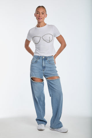 A person in the Rya Crystal T-Shirt - White and light blue jeans with knee cut-outs joined by chains in a bustier design stands against a plain white background with hands on hips.