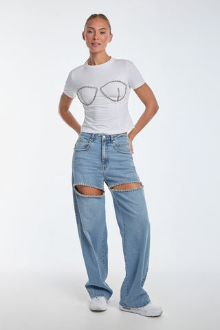 A person poses against a plain background in the Rya Crystal T-Shirt - White with chain details, light blue jeans featuring knee chain accents, and white sneakers. Hand-beaded crystal embellishments add a subtle sparkle as they smile confidently with hands on hips.