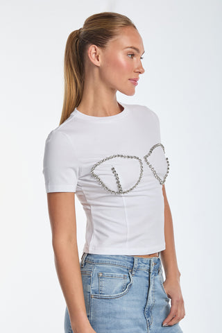 A person with long hair in a ponytail wears the Rya Crystal T-Shirt - White, featuring hand-beaded crystal embellishments forming gem-outline designs akin to a bustier. They pair it with blue jeans, standing against a plain white background and showing their side profile.