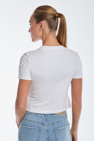 A person with a ponytail faces away, wearing the sleek white Rya Crystal T-Shirt with bustier design details and fitted blue jeans against a plain, light background.