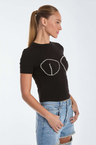 A woman wearing a black Rya Crystal T-Shirt, with pearl-like embellishments outlining a bustier design, stands against a white background. Her light hair is in a ponytail, and she pairs the top with denim shorts featuring similar decorations, facing sideways.