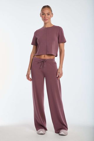 A woman models the Daphne Pant in Orchid, part of a mauve lounge set made from a soft modal blend. She wears a short-sleeve cropped top with wide-leg pants, exuding effortless style against a plain white background as she smiles barefoot, capturing the ultimate on-the-go look.