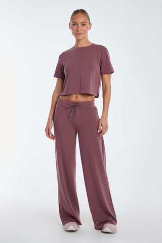 A person is wearing the Daphne Soft T-Shirt in orchid, styled in a cropped short-sleeve design with wide-leg pants. They stand on a light background, are wearing white sneakers, and have their hair tied back.