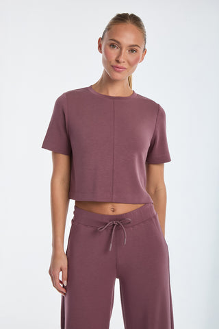 Wearing the Daphne Soft T-Shirt - Orchid in mauve and matching high-waisted pants, a person stands against a white background, smiling gently with sleek, pulled-back hair.