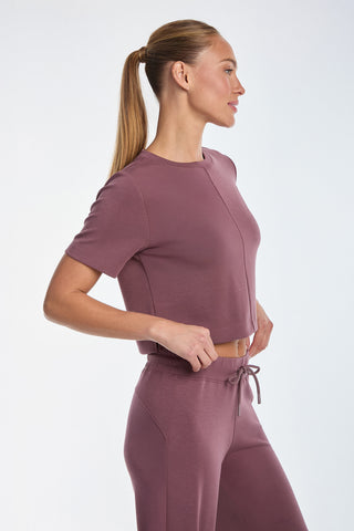 A woman with a ponytail in a fitted Daphne Soft T-Shirt - Orchid and matching pants, made of ultra-soft fabric, stands in profile against a plain background. She has her hands on her hips and is looking to her right.