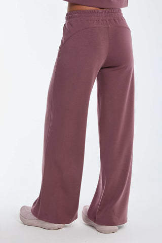 The image features a person in Daphne Pant - Orchid, loose-fitting mauve pants made from a soft modal blend for luxurious comfort, with an elastic waistband and pockets. Paired with white sneakers, they offer the ultimate on-the-go look against a plain white background.