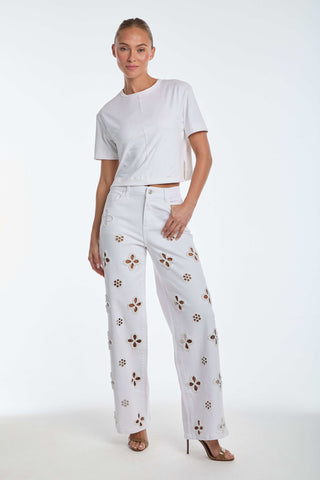 A person stands against a plain background, wearing a white short-sleeve top and Kaia Pearl High Rise Jean - White, featuring embroidered beaded flowers. They are paired with beige heeled sandals and have their hair pulled back.