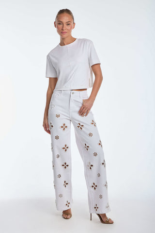 Person in a white short-sleeve crop top and Kaia Pearl High Rise Jean - White, featuring delicate embroidered beaded flowers. They pose on a plain white background, with one hand on their hip and the other relaxed by their side, wearing open-toe heels.