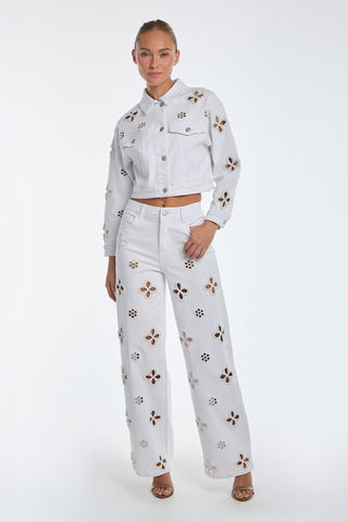 A person models the Kaia Pearl High Rise Jean in white paired with a matching denim jacket featuring embroidered beaded flowers, set against a plain background. They wear open-toed shoes and have their hair styled in an elegant updo.