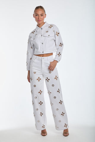 A person confidently wears a white denim jacket with Kaia Pearl High Rise Jeans, featuring embroidered beaded flower cutouts, paired with heeled sandals against a plain white background.